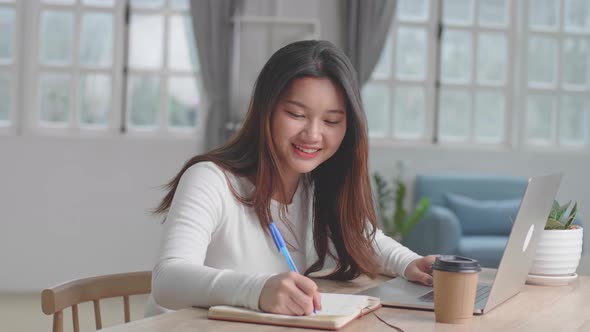 Happy Asian Girl Using Laptop Computer And Writing On Notebook At Home