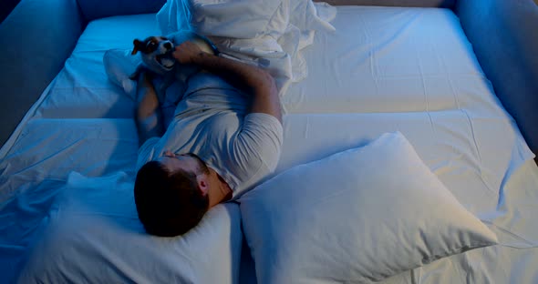 a Man Is Lying on a Sofa with White Bed Linen, Playing with His Dog and Putting the Dog's Tail in