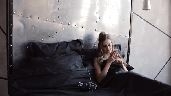 Gorgeous Blondie Checking Her Mobile Phone Sitting in Bed Under Black Blanket