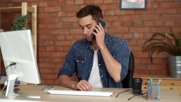 Front View of Happy Young Business Man Working Typing on Computer Taking Incoming Call on Mobile