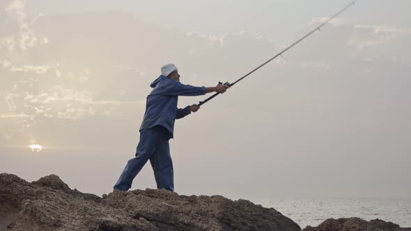 Old fisherman standing on sea side rocks and fishing against the sunset