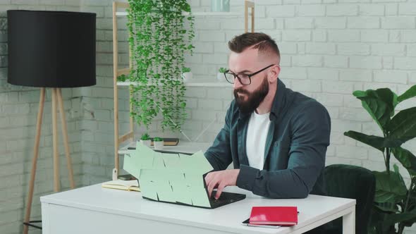 Bearded Man Types at the Laptop and Write Notice on the Paper Notebook