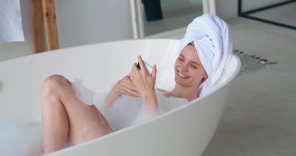Young Charming Woman with a White Terry Towel on Head Lying Naked in Bathtub Smiles Using Mobile
