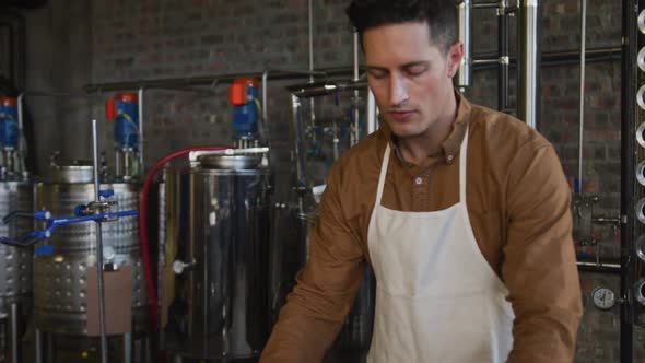 Portrait of serious caucasian man working at gin distillery, using equipment and looking to camera