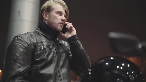 A Young Man Wearing a Leather Jacket Sits on His Motorcycle at the Sunset and Speaks on the Phone at