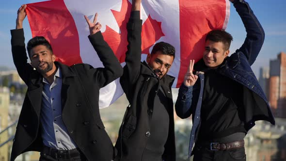 Three Positive Men with Canadian Flag Looking at Camera Gesturing Peace Standing on Rooftop