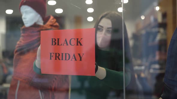 Young Brunette Woman in Covid-19 Face Mask Hanging Black Friday Sales Announcement on Shop Window