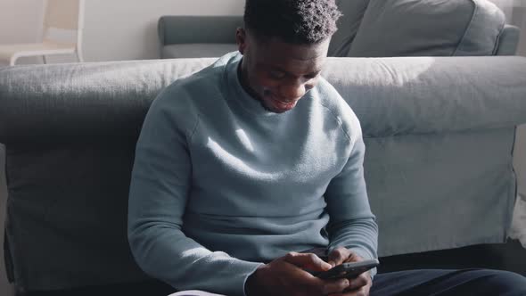 African American Black Man Sitting on the Floor in a Cozy Room at Home Using His Cell Phone