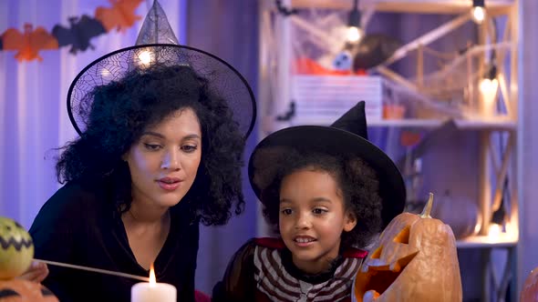 Little Girl and Woman African American in Witch Hat Light Candle in Pumpkin with Carved Smiling Face