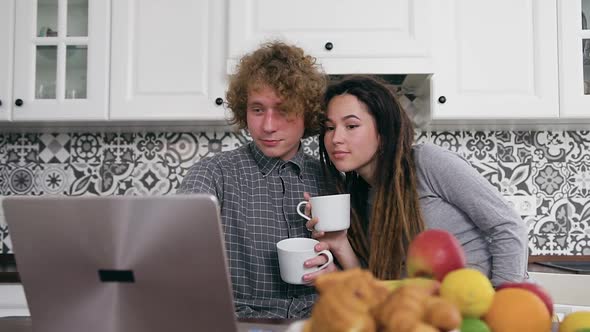 Pregnant Couple Sitting Near Kitchen Table, Drinking Tea and Watching on Laptop Screen