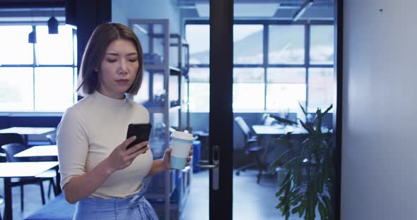 Asian businesswoman walking using smartphone holding cup of coffee in modern office