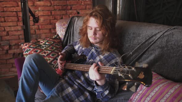 Portrait of Young Man Musician with Long Red Hair Playing Bass Guitar at Home Sitting on Sofa Slow