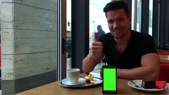 A Man Sits at a Table in a Cafe and Gesticulates Excitedly Toward the Smartphone with a Green Screen