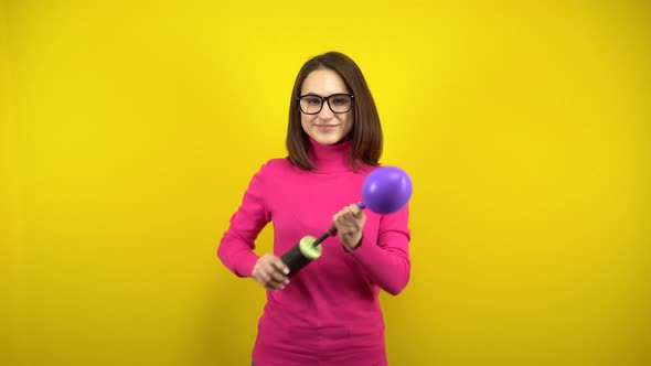 A Young Woman Inflates a Purple Balloon with a Pump on a Yellow Background. Girl in a Pink