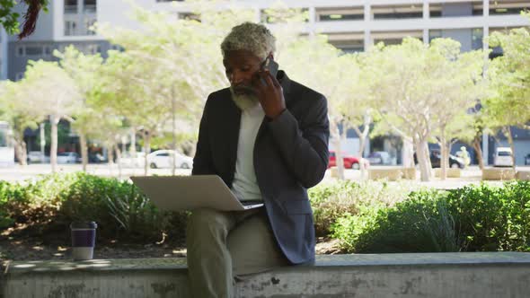 African american senior man talking on smartphone while using laptop in corporate park