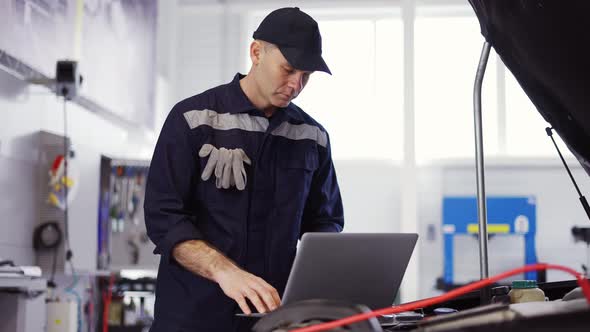 Portrait of Mechanic Uses a Laptop While Conducting Diagnostics Test on Engine Slow Motion