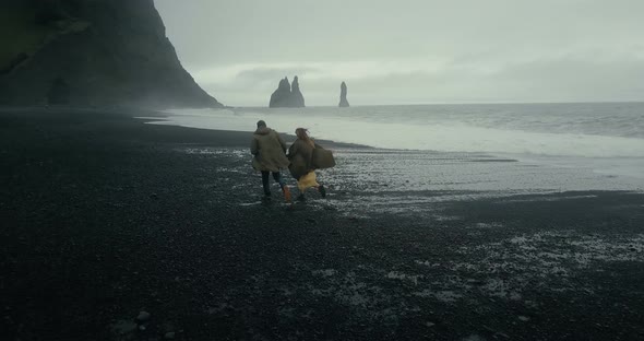 Back Aerial View of Young Hipster Couple Running on the Black Volcanic Beach Near the Troll Toes