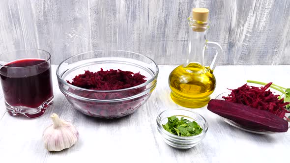 A glass of beet juice, grated beetroot in a salad bowl, garlic, parsley, and a bottle of olive oil
