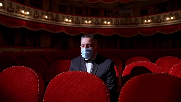 Visitor in suit and medical mask sitting in comfortable chairs alone on dark theater background.