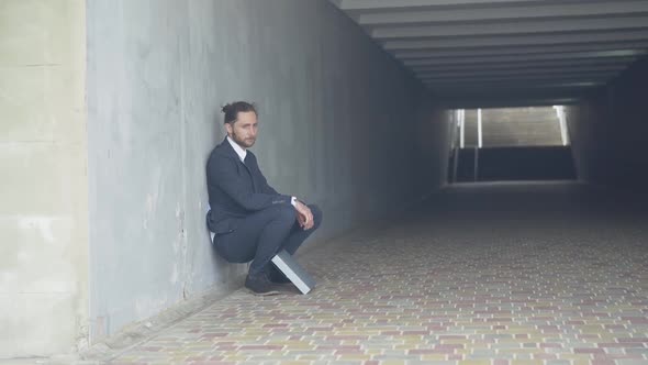 Wide Shot of Depressed Man in Suit Sitting Against the Wall in Underground Crossing. Frustrated