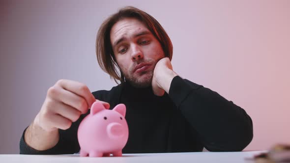 Young Upset Man Putting Small Amount of Coins in the Piggy Bank