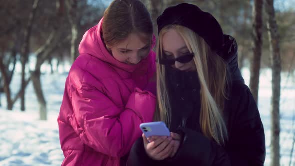 Two Trendy Teenage Smiling Girls in Pink and Black Jackets Look at Photo on Smartphone Together in