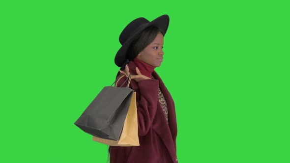 Pretty African American Fashion Girl in Coat and Black Hat Strolling with Shopping Bags on a Green