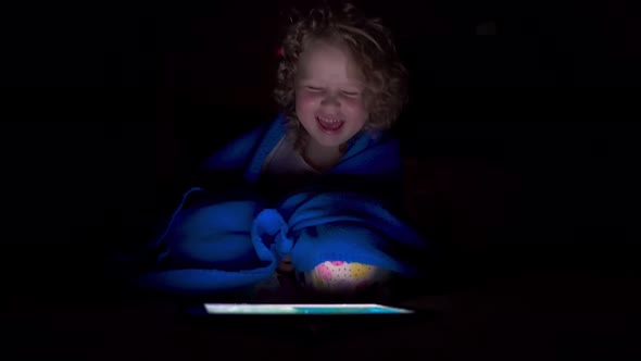 Little girl sits in bed under the blanket before going to bed and makes a video call