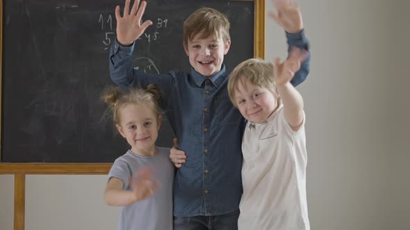 Cheerful Caucasian Friends or Siblings Waving Looking at Camera Standing in Classroom at Chalkboard