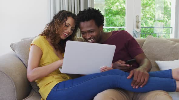 Happy biracial couple sitting on sofa with laptop and laughing