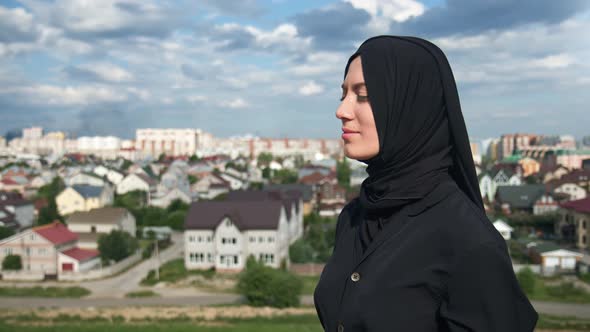 Pleasant Muslim Woman in Traditional Black Headscarf Standing on Mountain Peak Over Islamic City