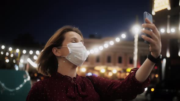 Beautiful woman in medical mask using phone making a video call smiling and talking with friend.