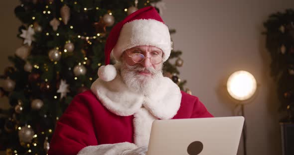 Cheerful Elderly Bearded Santa Claus Sitting at a Table Drinking Hot Drink While Working on Laptop