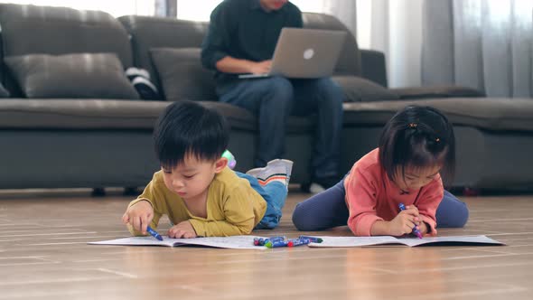 Cute Little Boy And Girl Lying On The Floor And Drawing While Their Father Working On Laptop