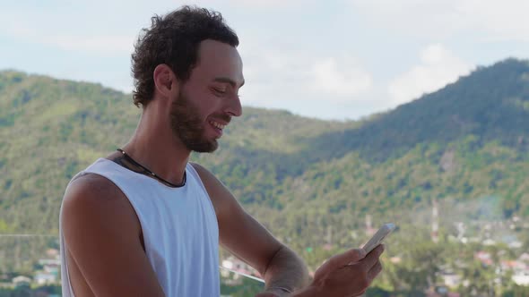 Charming Young Caucasian Man Dressed in a Casual White Tshirt Stands on His Hotel Balcony