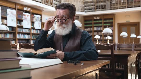 Old Bearded Man with Wrinkled Face which Putting on His Glasses and Reading Book in Library