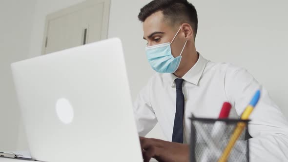 Handsome Male Student in Medical Mask Thinking When Typing on a Laptop at Table