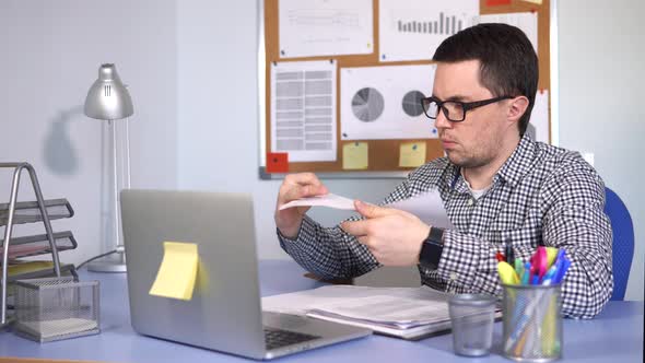 Busy Man Fills Out Paper Forms in the Workplace and Looks at the Laptop Monitor