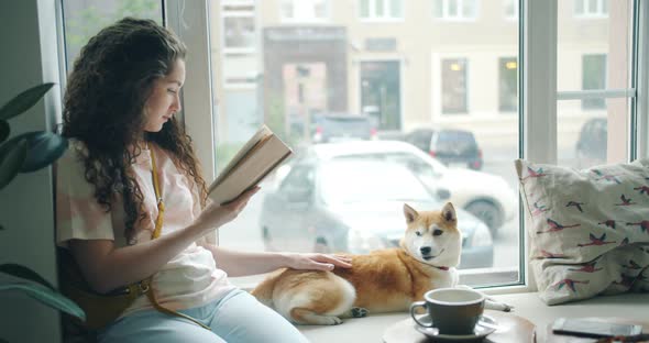 Attractive Girl Dog Owner Reading Book and Stroking Pet on Window Sill in Cafe