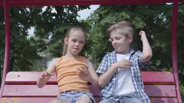 Adorable Boy and Cute Girl Sitting on the Swing Close Up in the Park, Dancing, Having Fun Looking in