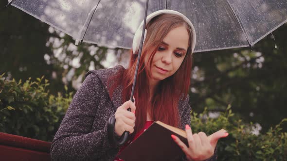 Young Beautiful Woman in Headphones with Transparent Umbrella Reading Book Sitting on Bench in City