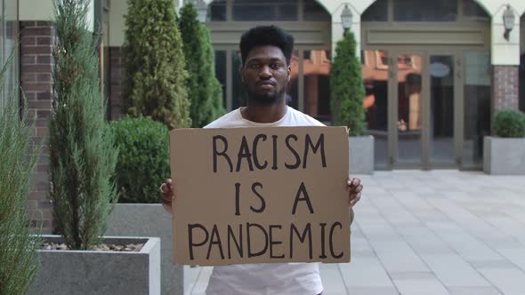 A Young African American Man Stands with a Cardboard Poster RACISM IS A PANDEMIC in a Public Outdoor