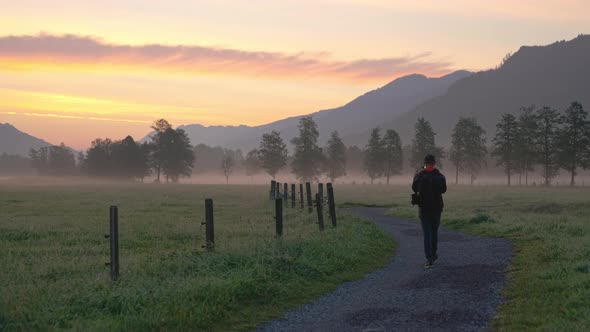 Man Walking Along Path Towards Misty Landscape At Dawn