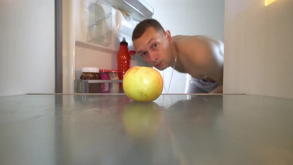 young man opens refrigerator and sees empty shelves. One apple is lying.