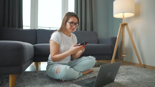 Casually Dressed Woman is Sitting on Carpet with Laptop and Smartphone and Working in Cozy Room