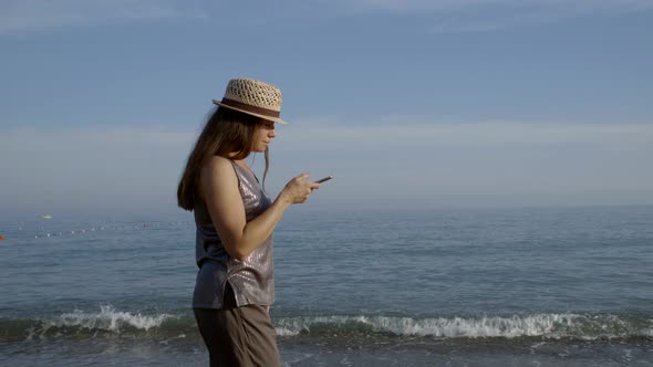 Vacationer Woman with Straw Hat on Head Is Walking Near Sea and Looking at Screen of Smartphone