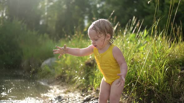 Little Funny Cute Blonde Girl Child Toddler in Yellow Wet Bodysuit Playing By the Lake Waterside