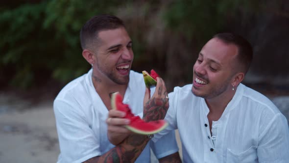 Diverse Gay Male Couple Eating Watermelon Together and Talking Sitting on the Rocky Beach