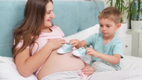 Smiling Pregnant Woman Looking on Her Little Son Playing with Baby Boots on Her Big Belly