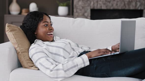 African Emotional Girl American Woman Looking at Laptop Lying on Sofa at Home Feels Delighted